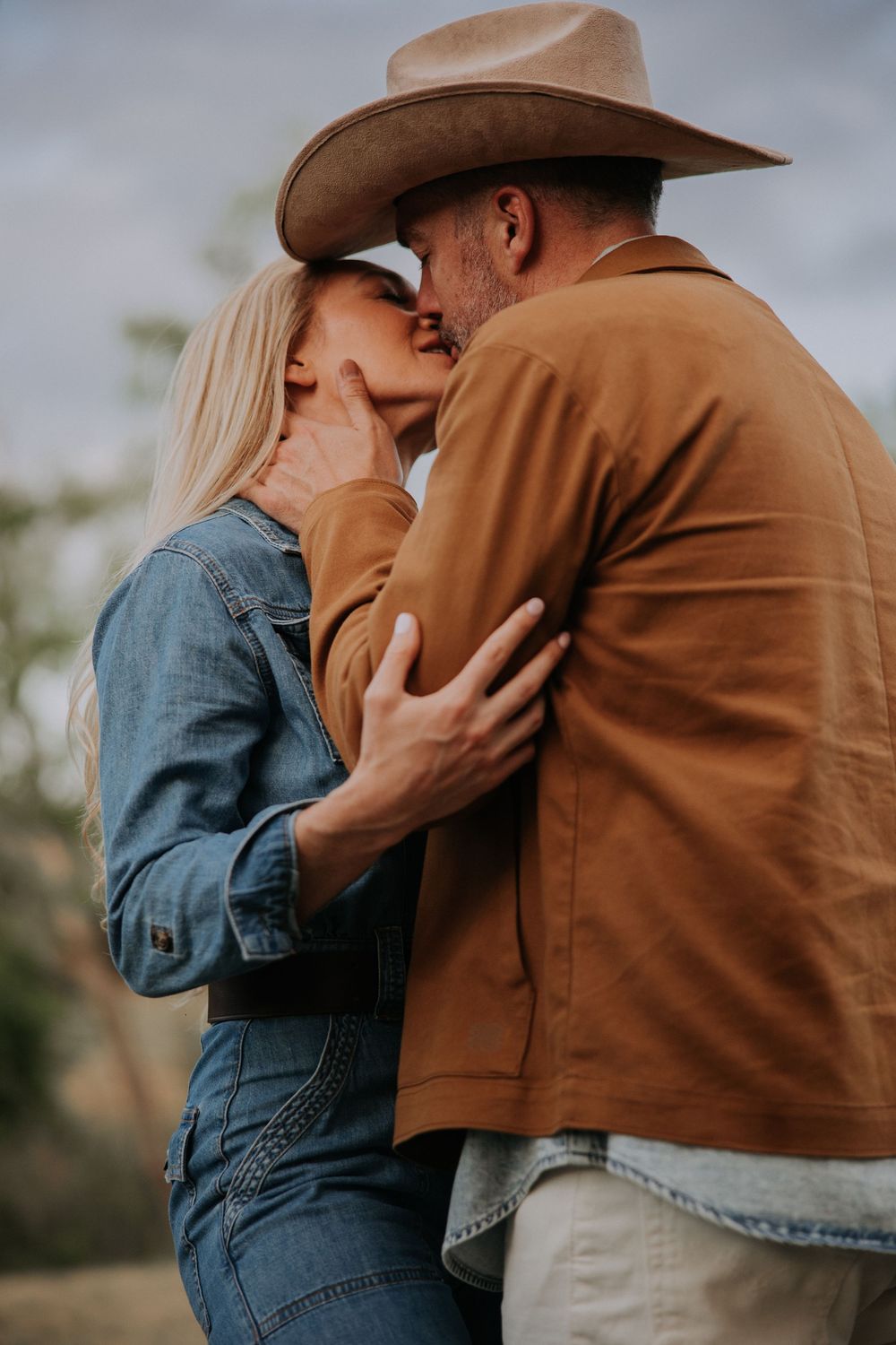 Couple sharing a romantic kiss outdoors, man wearing a cowboy hat and brown jacket, woman in a denim outfit.