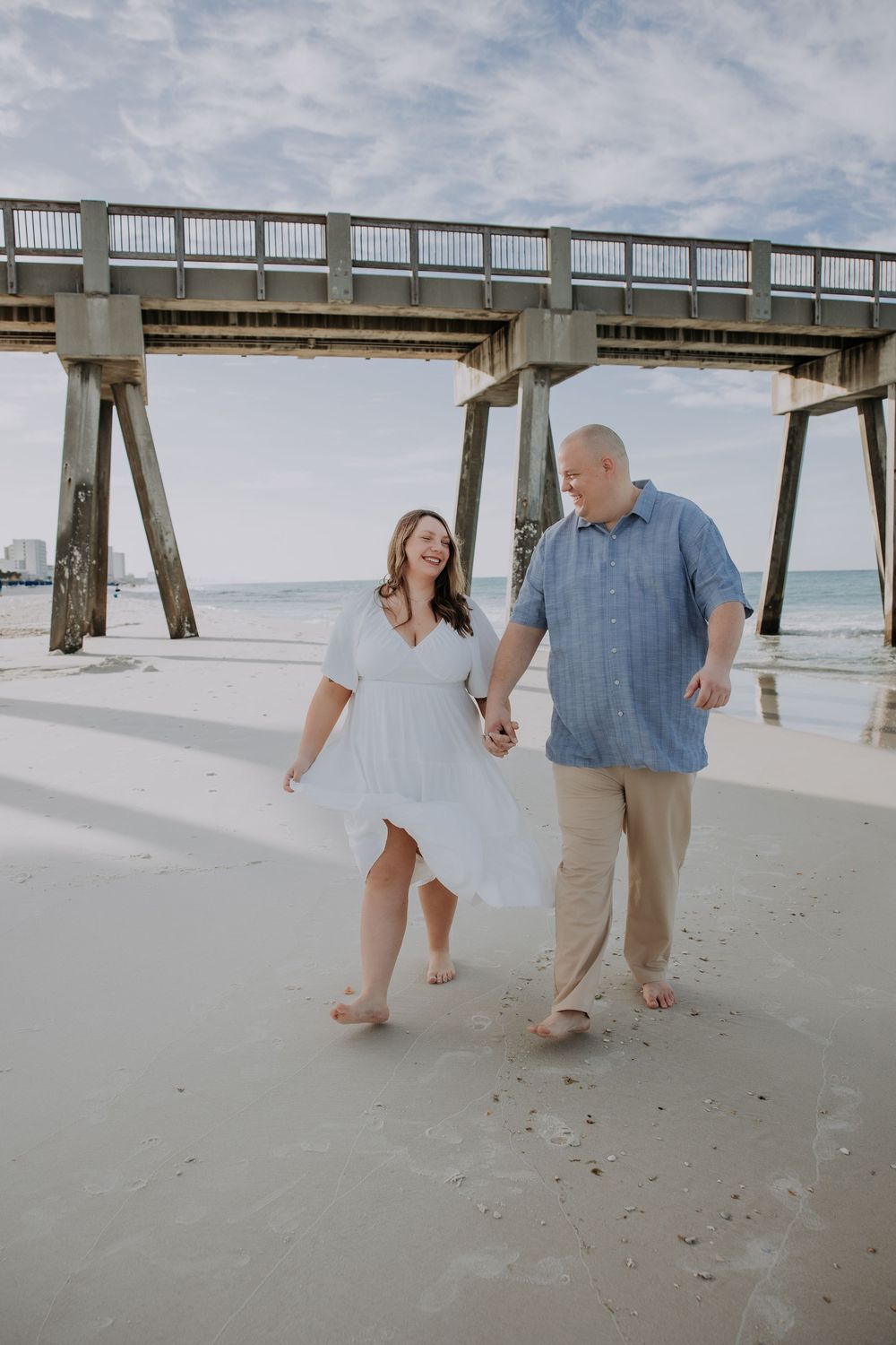 Couple holding hands and walking barefoot on sandy beach near a pier, woman in white dress, man in blue shirt.