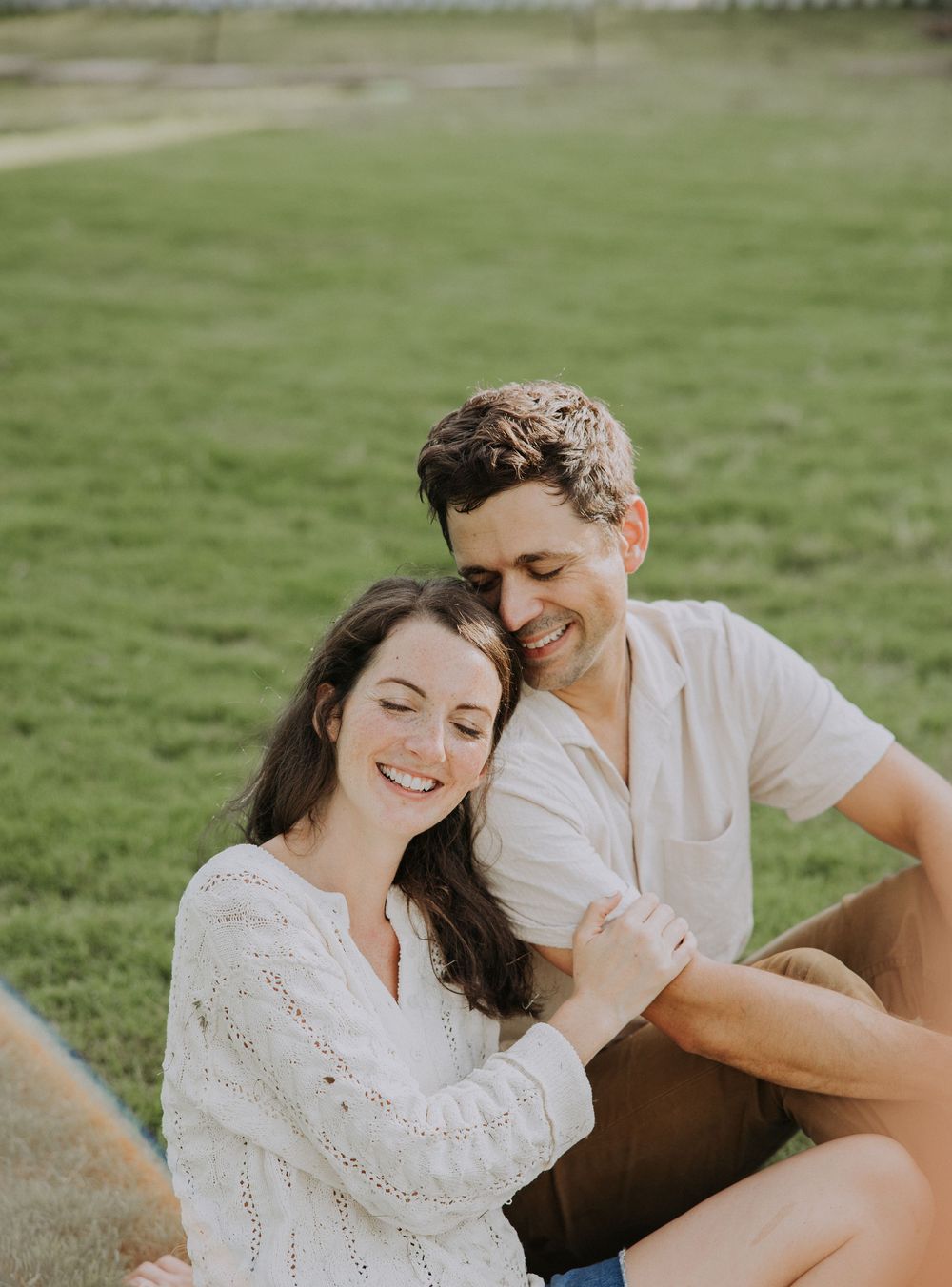 A smiling couple sits together on grass, the woman in a white sweater leaning into the man in a cream shirt.