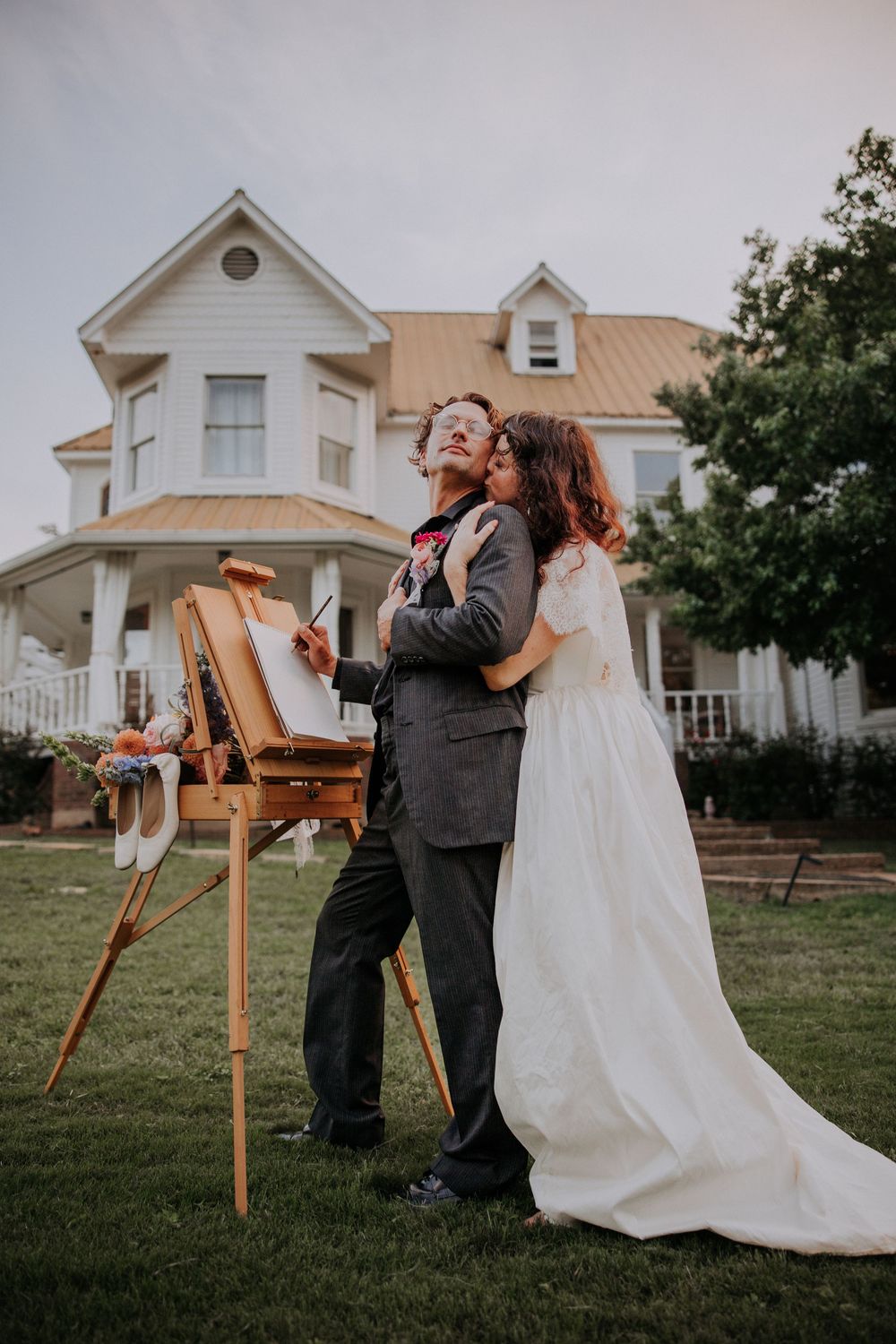 Bride in white gown embraces groom painting at easel outside elegant white farmhouse during outdoor wedding.