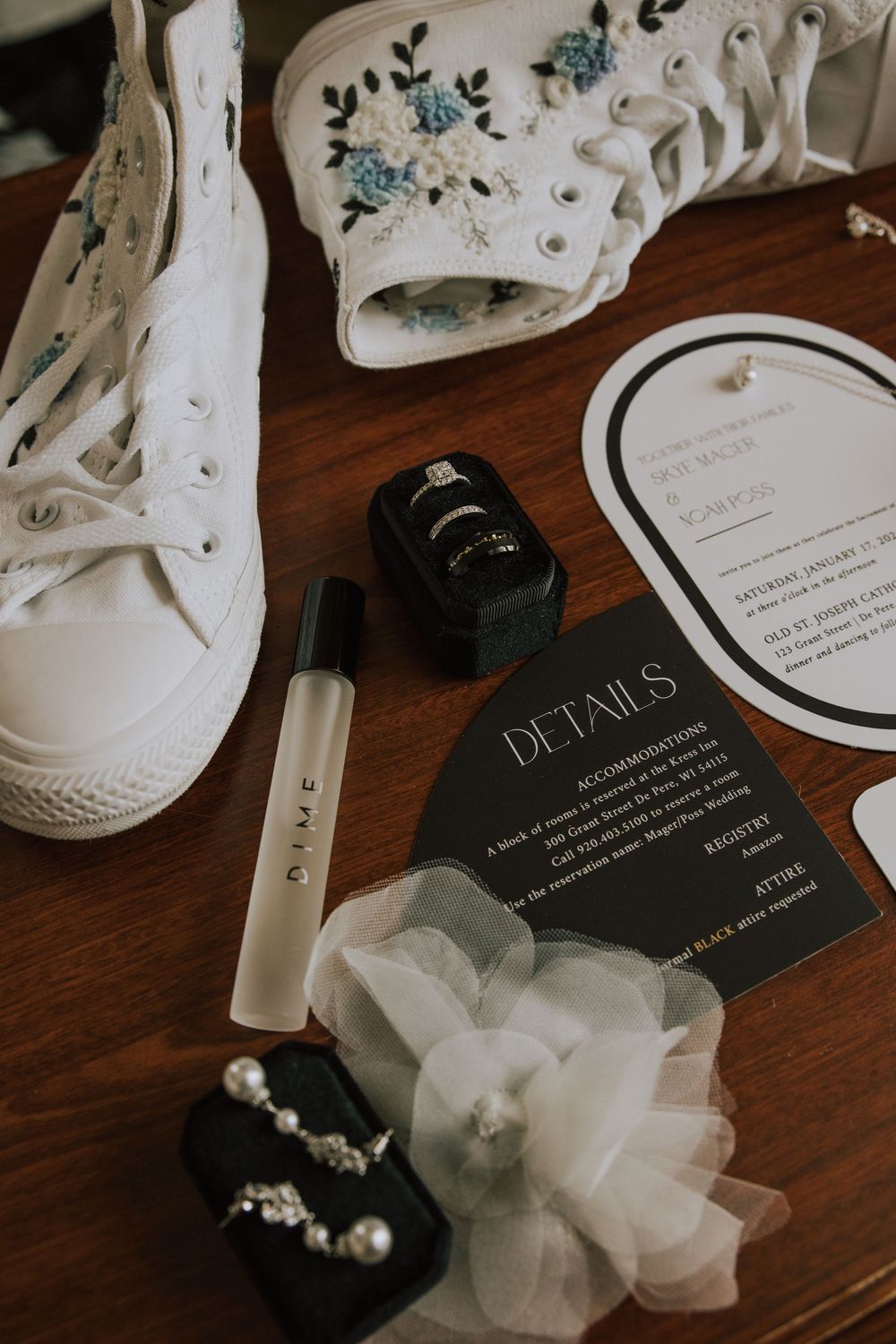 Wedding flat lay featuring floral Converse sneakers, ring box, perfume, pearl earrings, and elegant black details card on wood table.