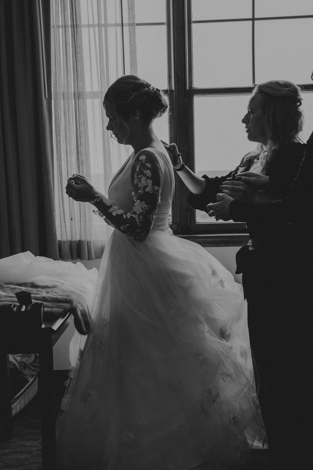 Black and white photo of a bride in a lace gown being helped by a woman near a bright window.