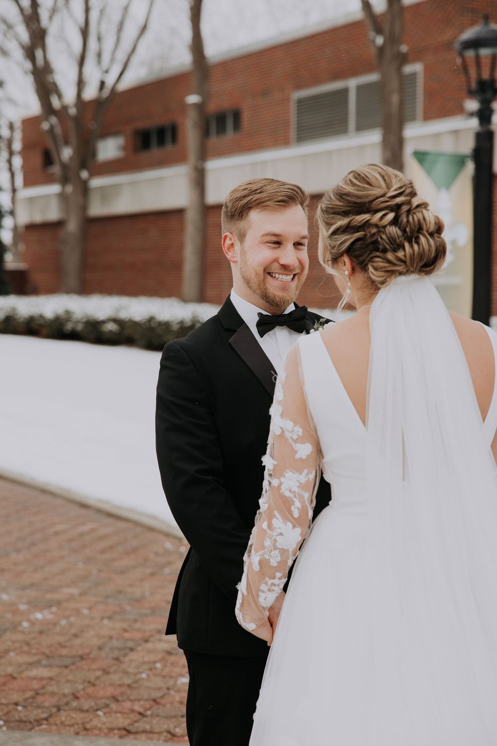 Smiling groom in black tuxedo holds hands with bride in lace wedding dress and veil outdoors in winter.