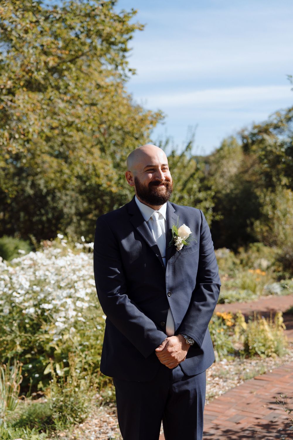 Wedding party member in navy suit standing in garden with white daisies on sunny day.