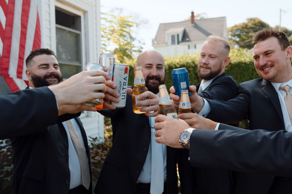 Groomsmen in dark suits toasting with drinks during wedding celebrations.