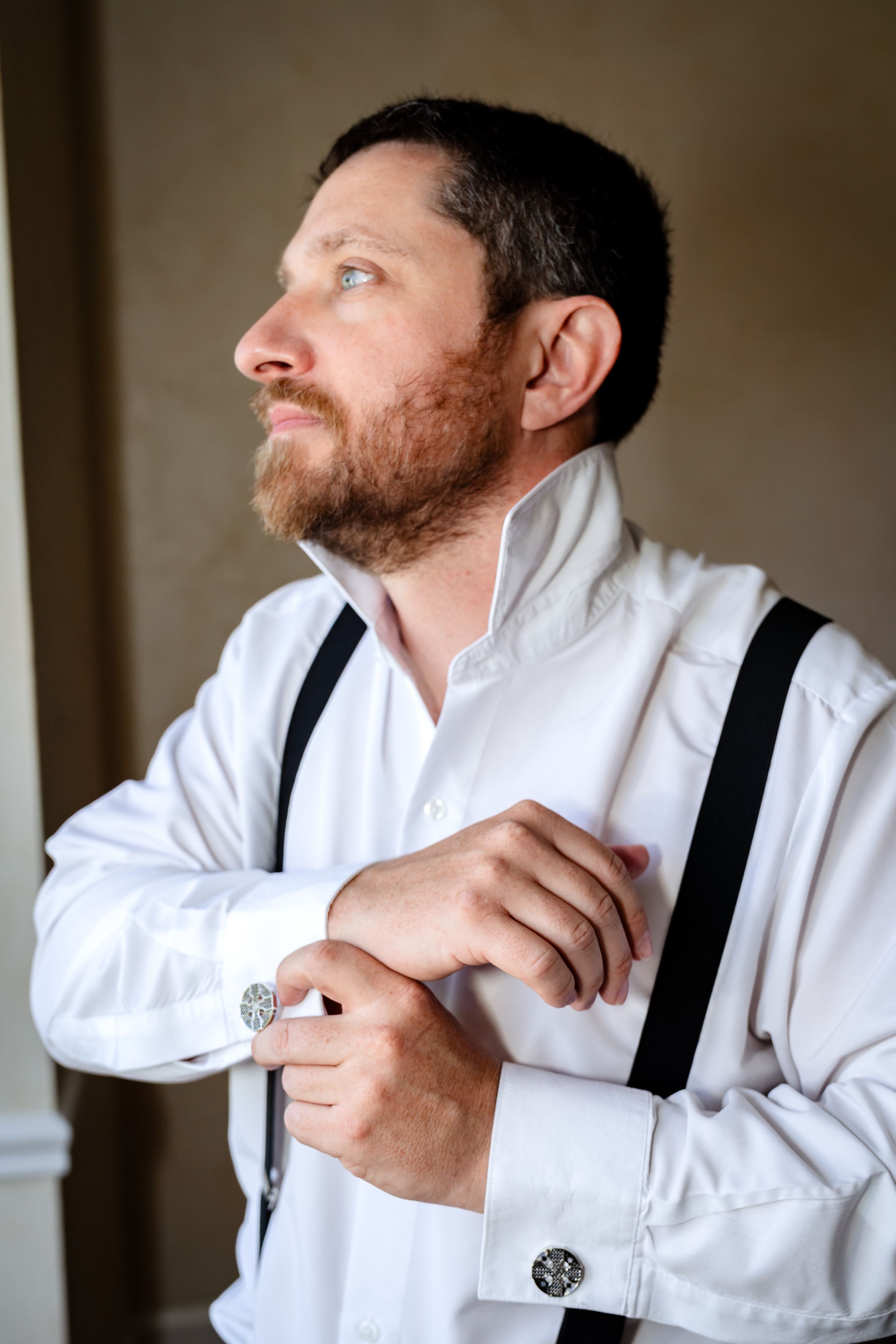 Groom adjusting white dress shirt and suspenders while getting ready.
