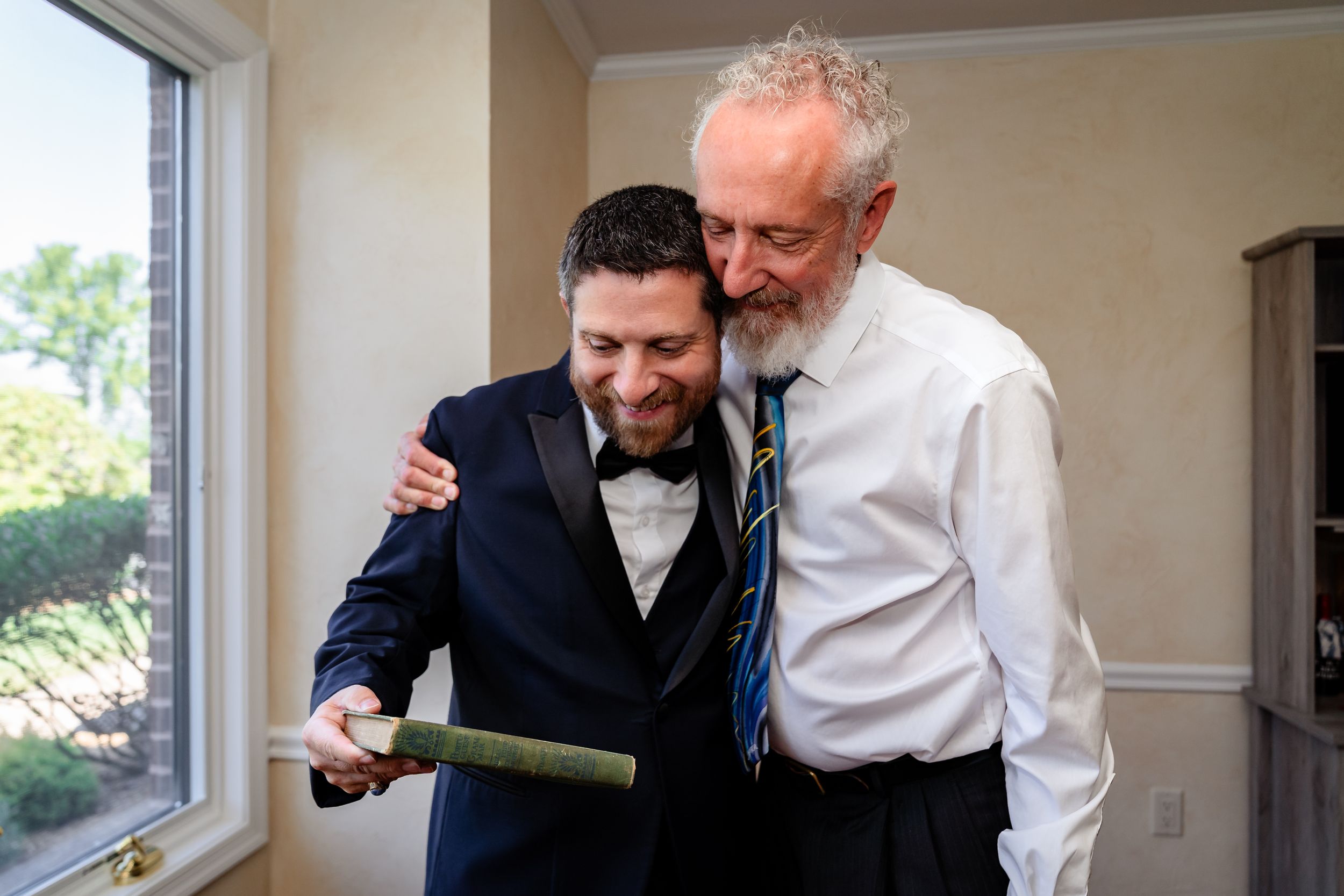 Groomsmen share a moment looking at something together by a bright window.