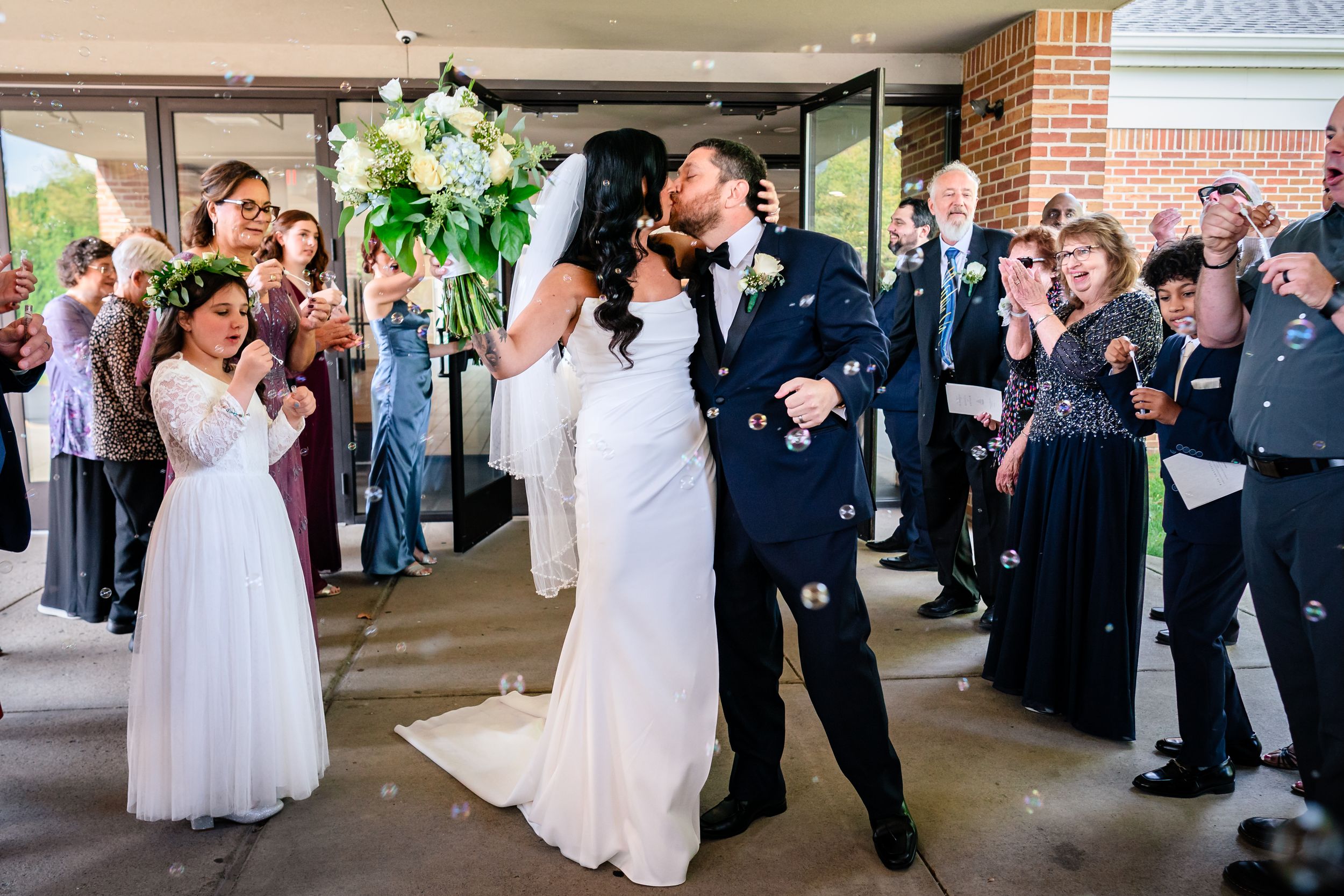 Wedding celebration with guests gathered under covered outdoor area near brick building.