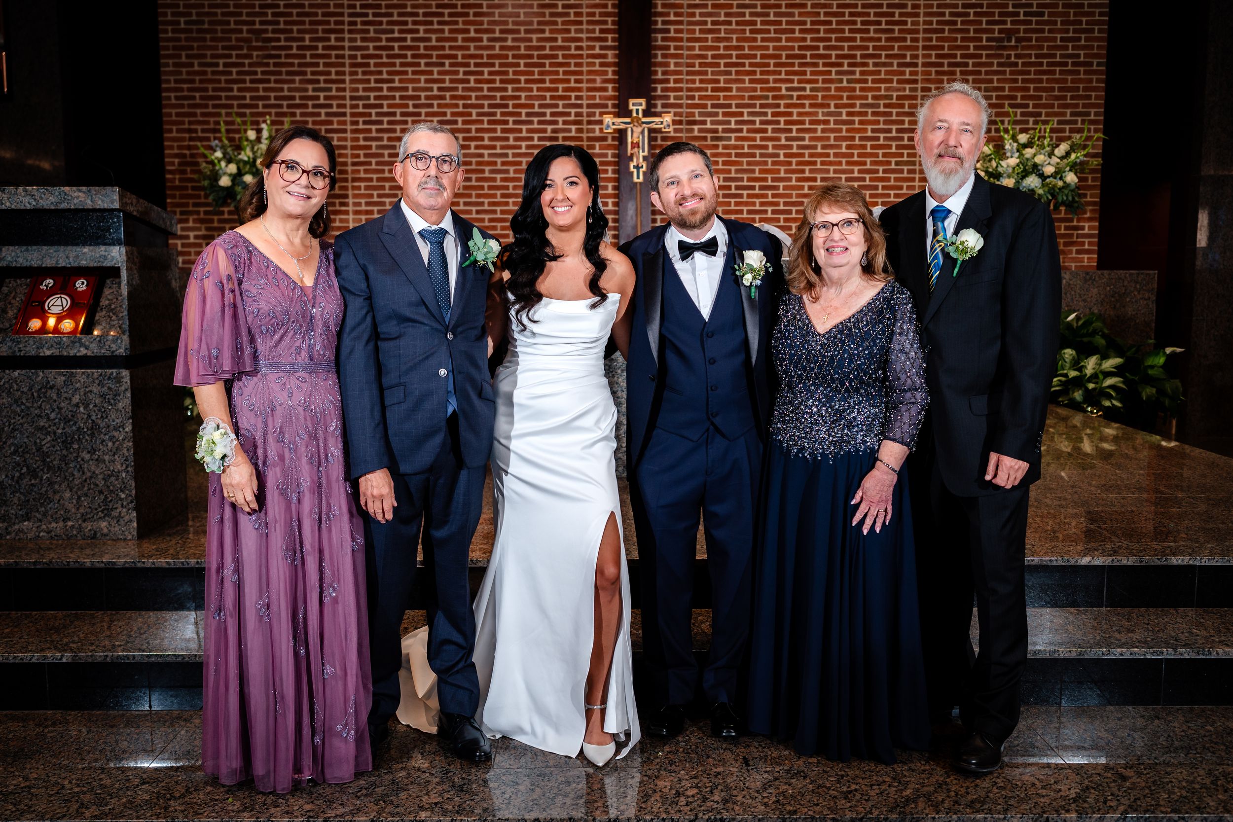 Formal wedding party group photo against brick wall with floral arrangements.