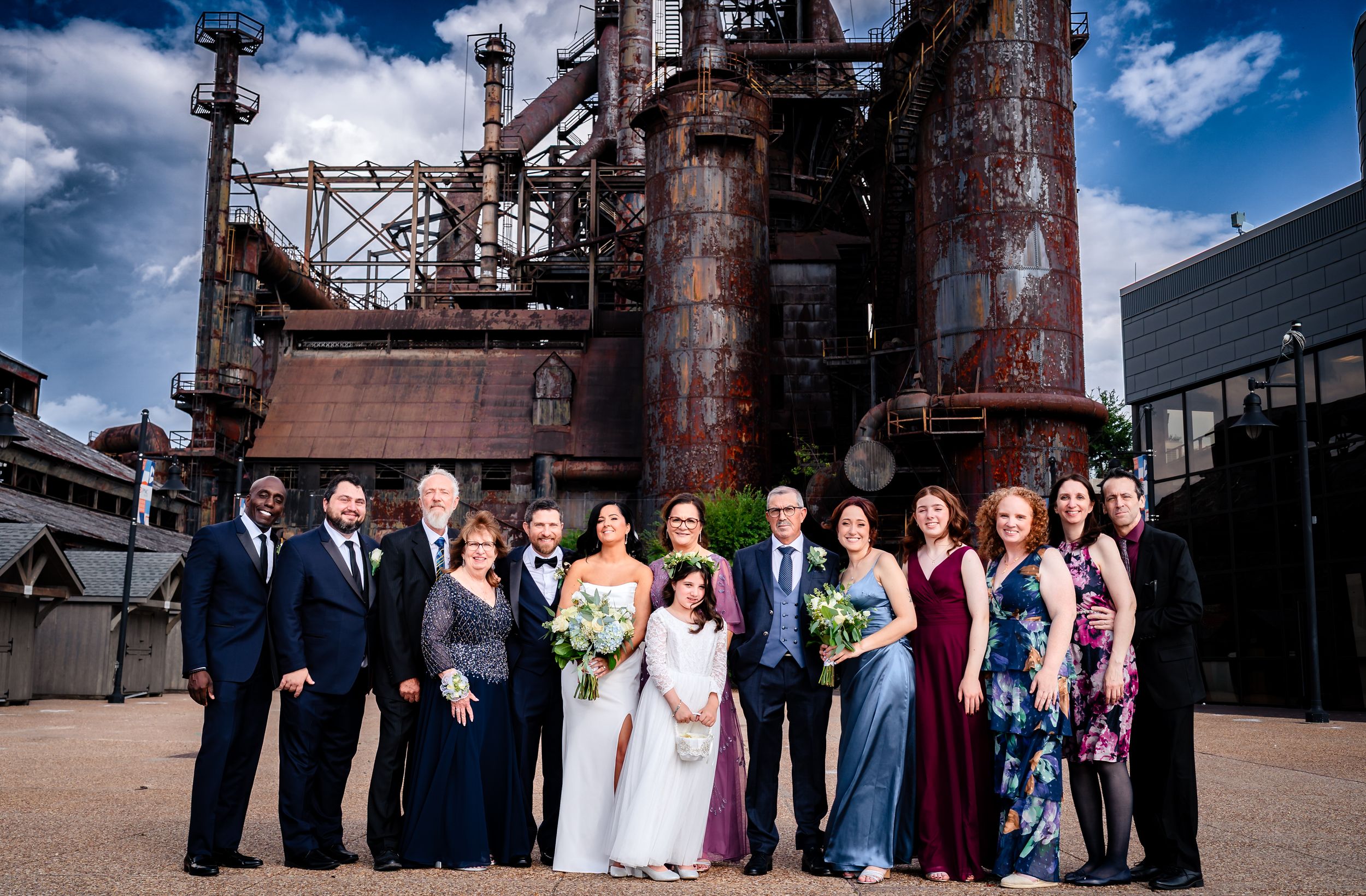 Wedding party group photos taken against industrial steel works backdrop under blue sky.