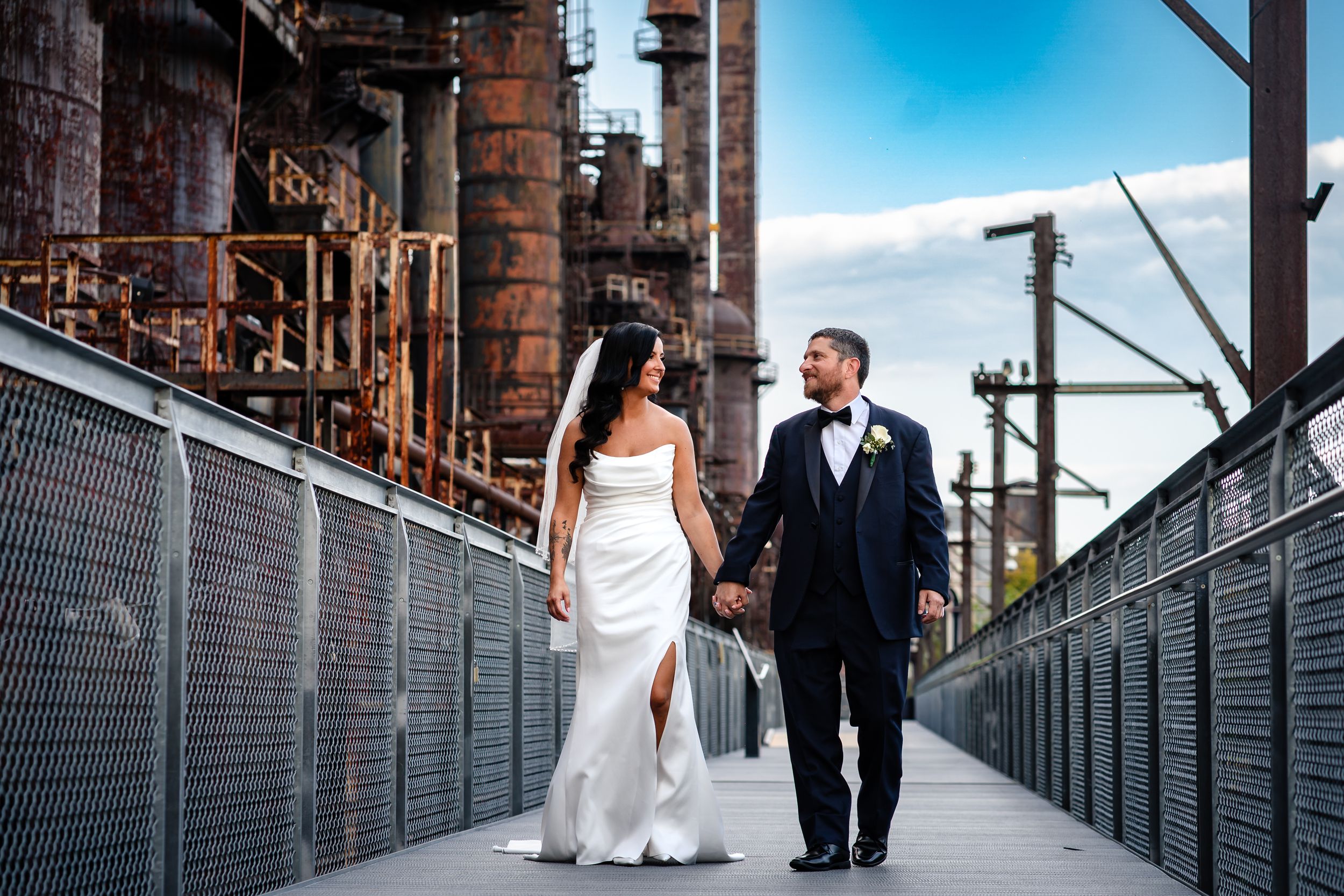 A couple walks along a metal bridge walkway against a bright blue sky.