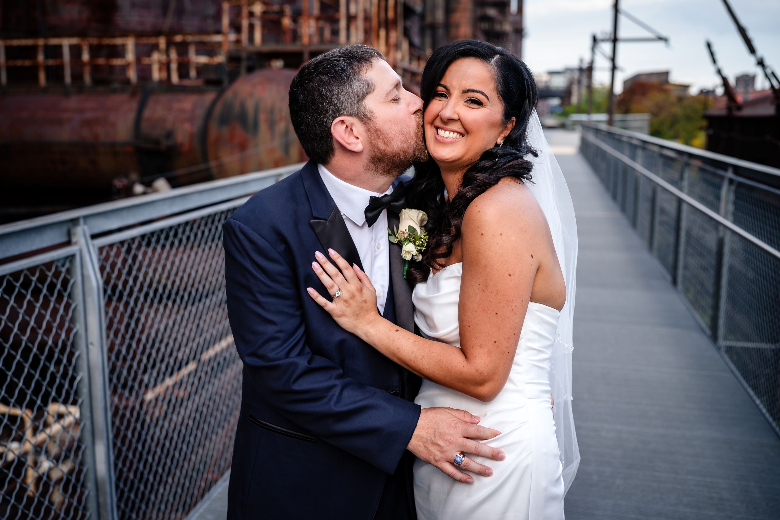 A couple embraces on an industrial walkway with urban architecture in the background.
