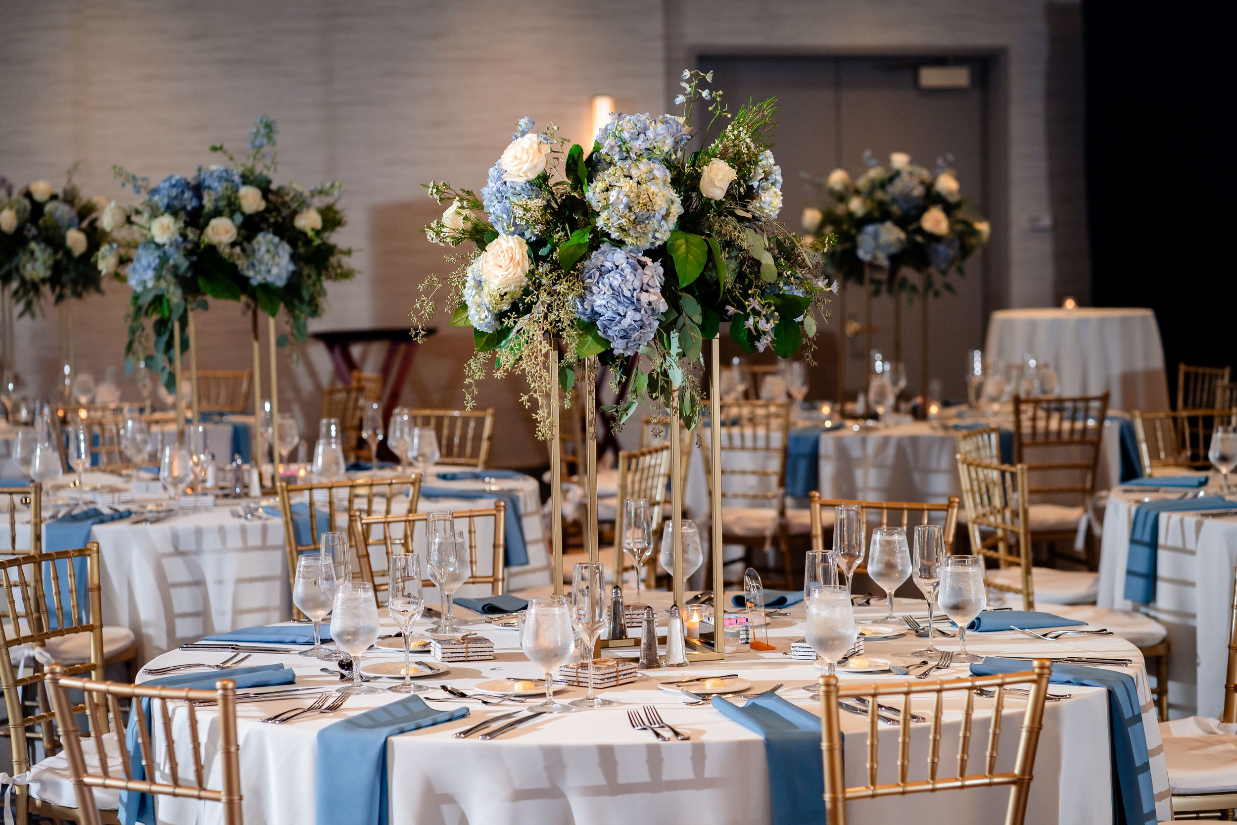An elegant wedding reception setup with blue and white floral centerpieces and gold chairs.
