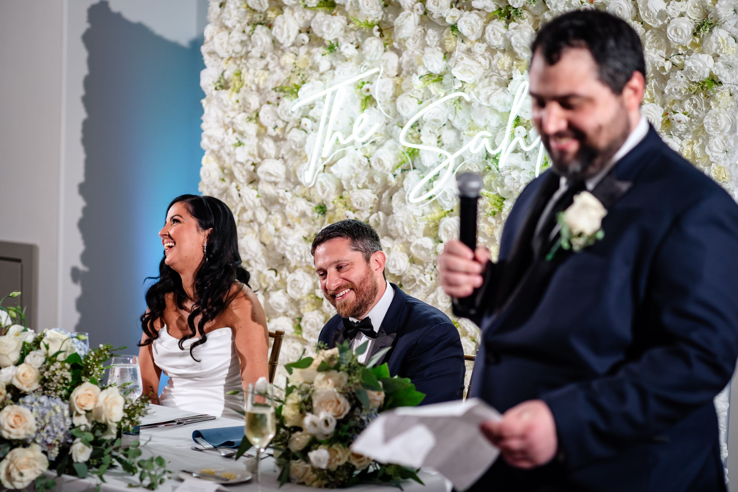 Wedding party members share joyful moments against a white floral backdrop.