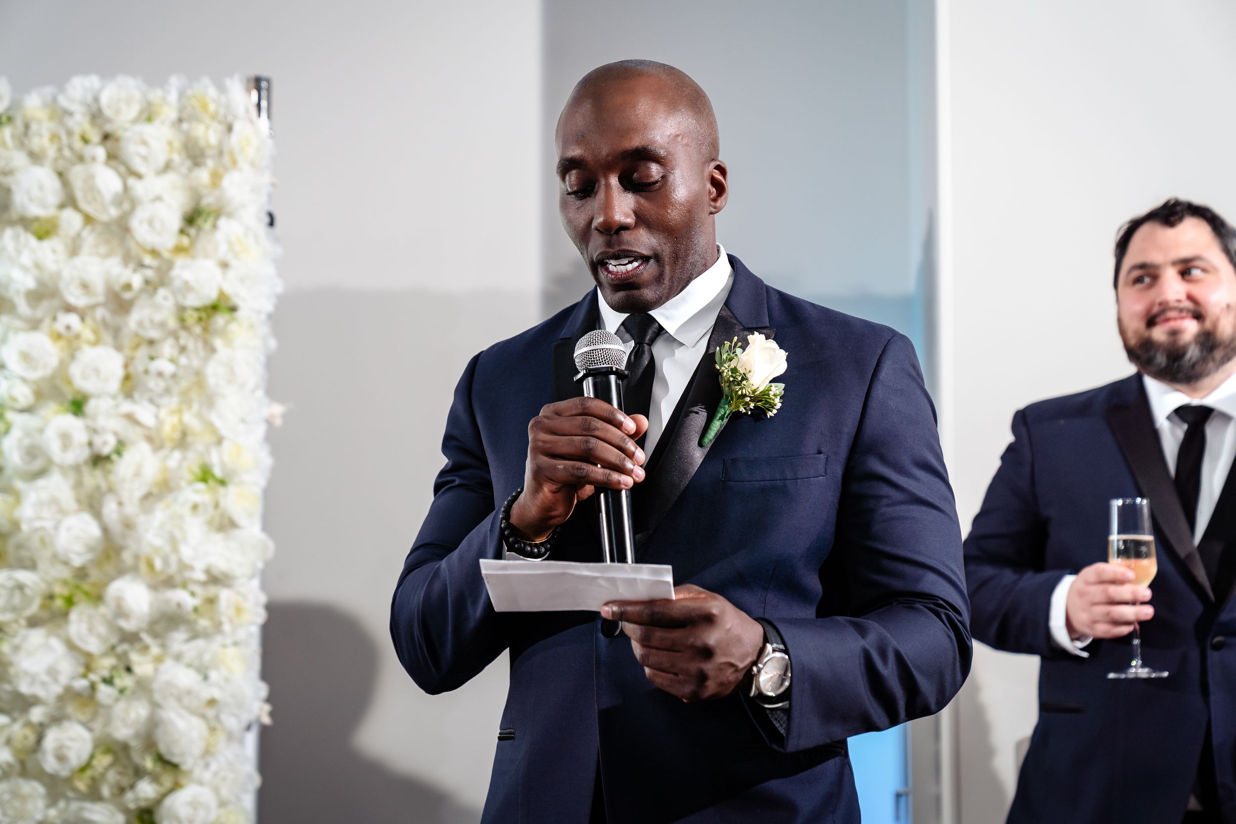 Wedding party members give toasts and speeches in front of an elegant white floral backdrop.