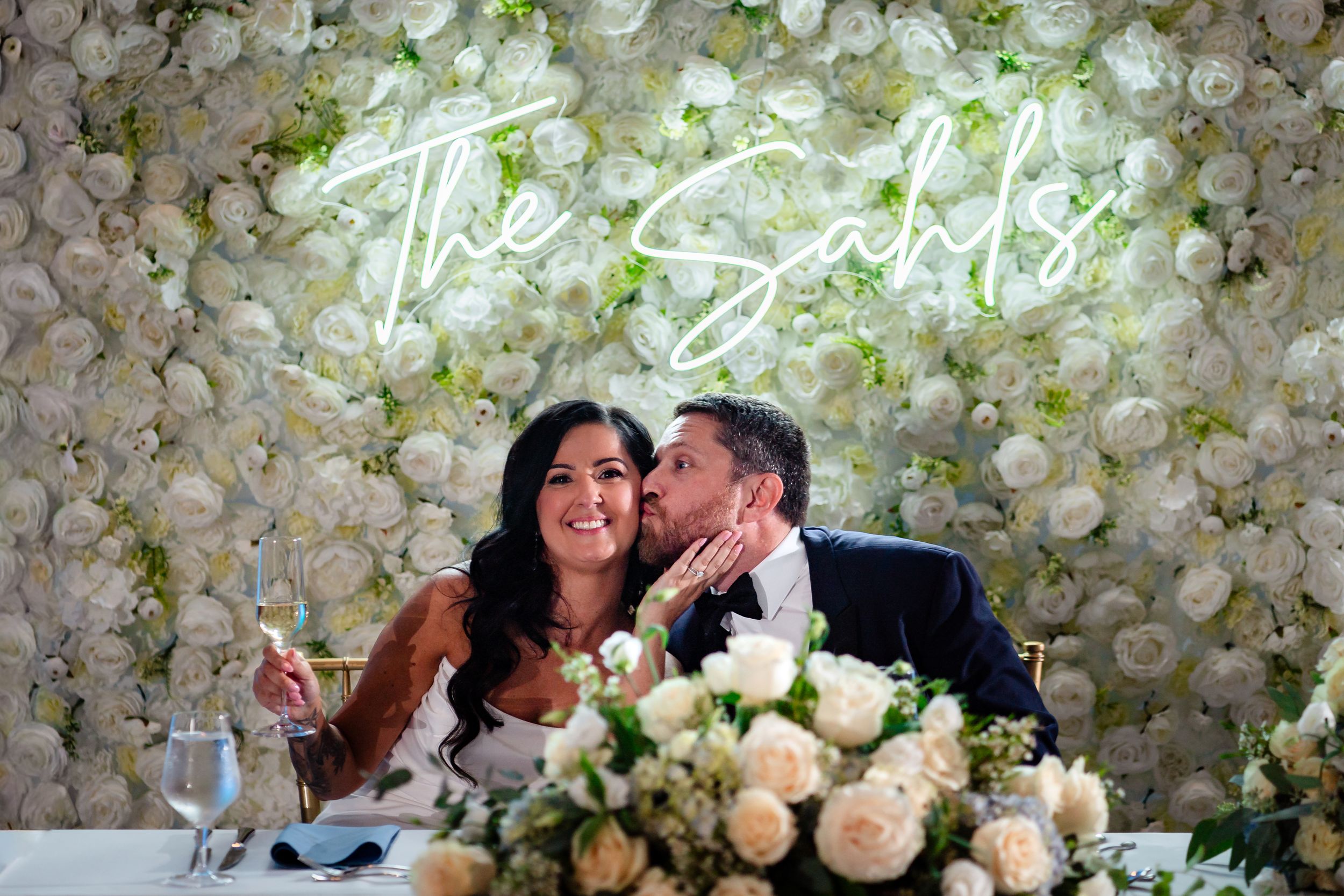 Guests enjoy emotional wedding speeches in front of a romantic white flower wall display.