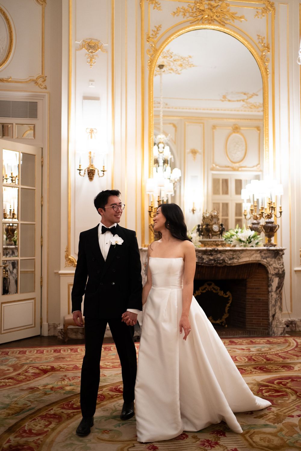 A couple in formal wedding attire walks through an elegant ballroom with ornate gold decor and crystal chandeliers.