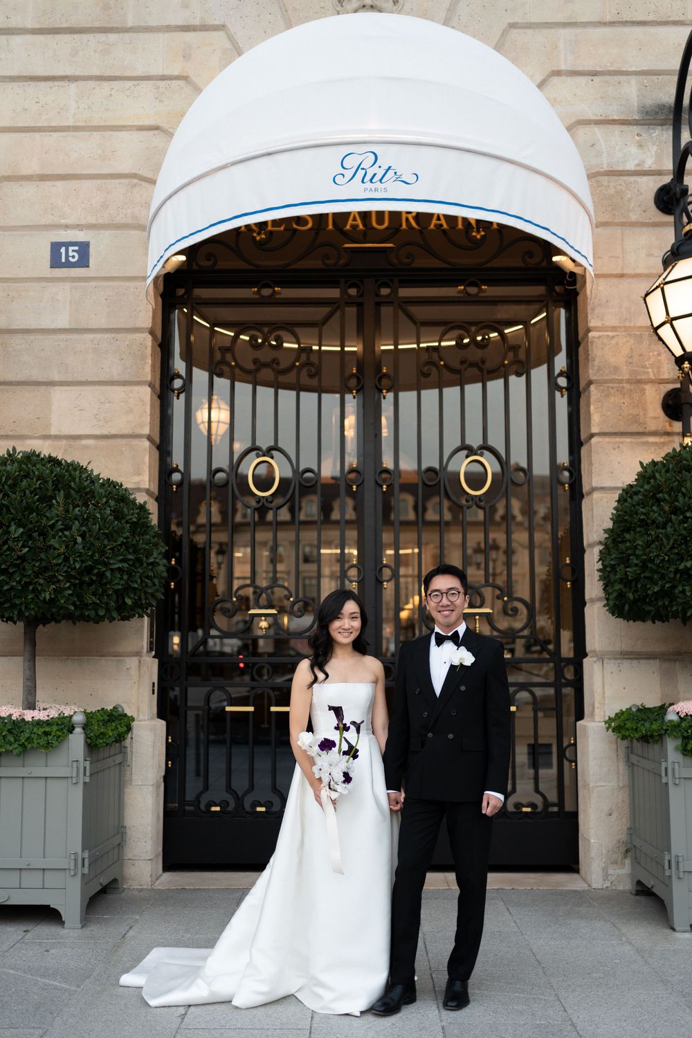 A wedding couple poses in front of an elegant stone building entrance with potted plants and ornate iron gates.