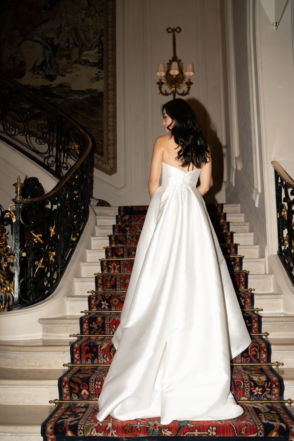 A flowing white wedding gown cascades down an elegant staircase adorned with a red patterned carpet. Ritz Paris wedding.