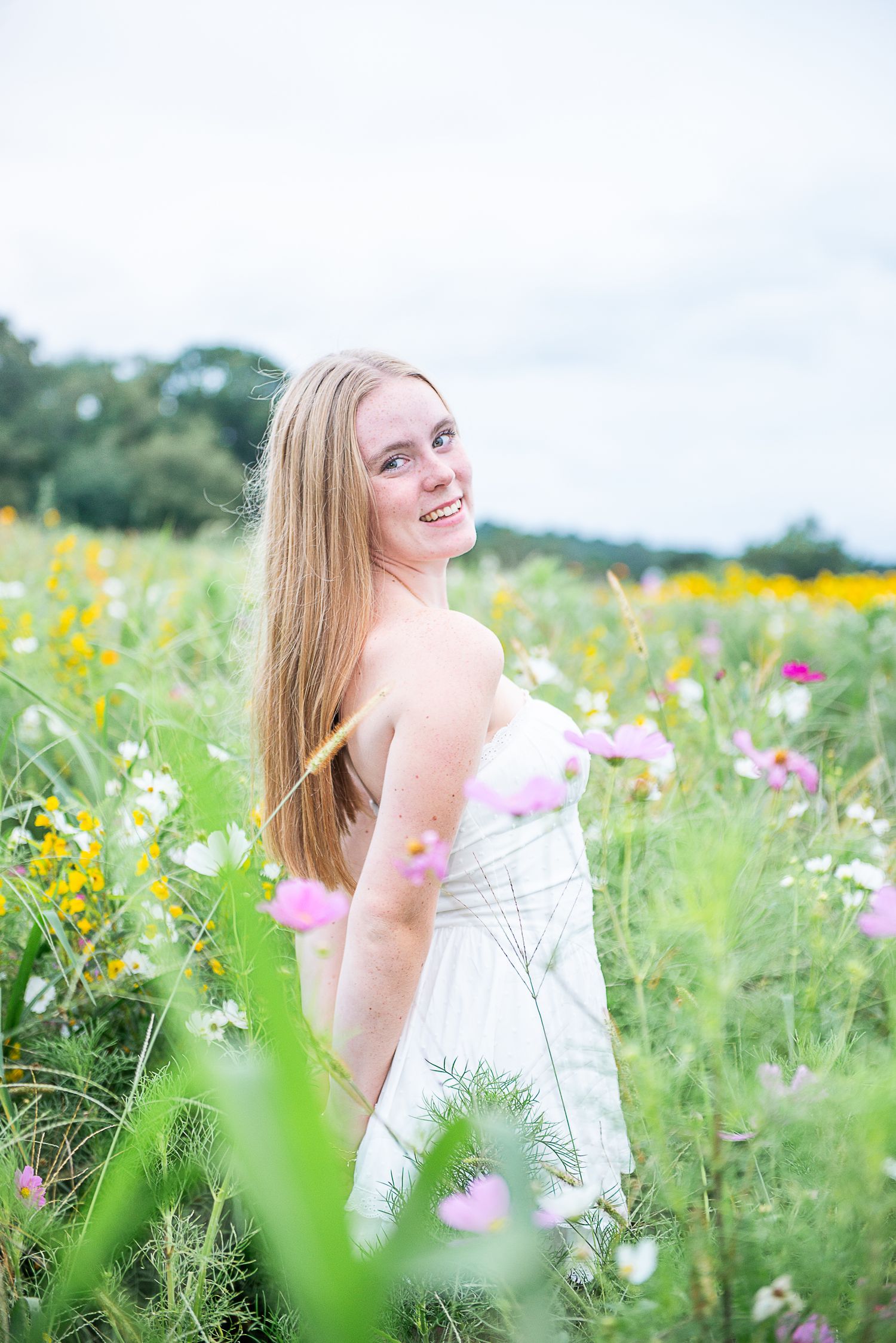 A person in a white dress poses joyfully in a vibrant summer wildflower meadow with cosmos and yellow blooms.
