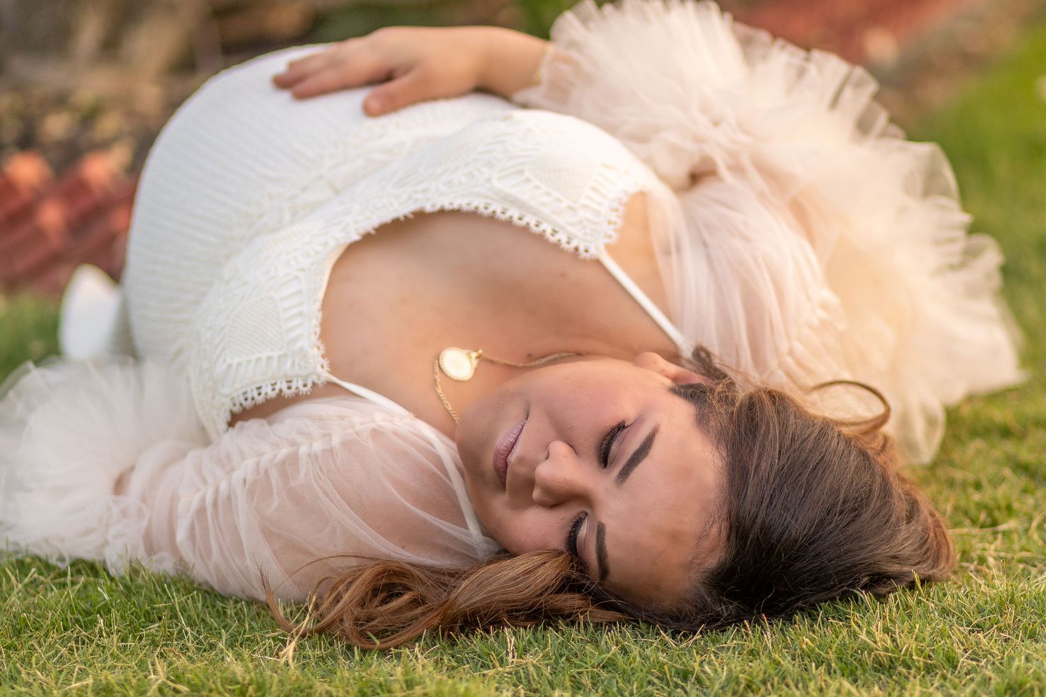 Expectant mother in white lace dress lying on grass during maternity photoshoot.