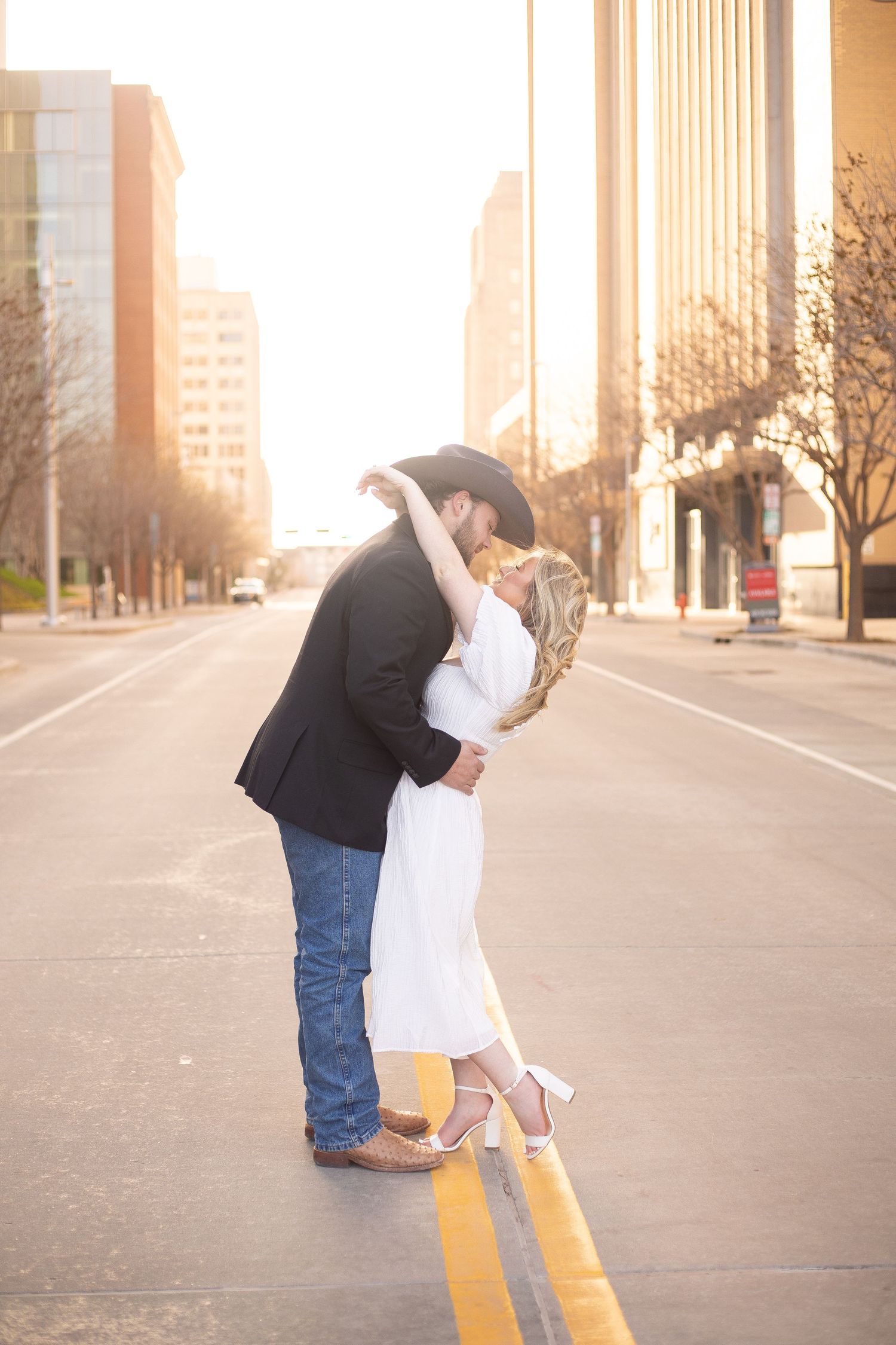 A romantic couple shares a kiss on a city street at sunset with urban buildings in the background.