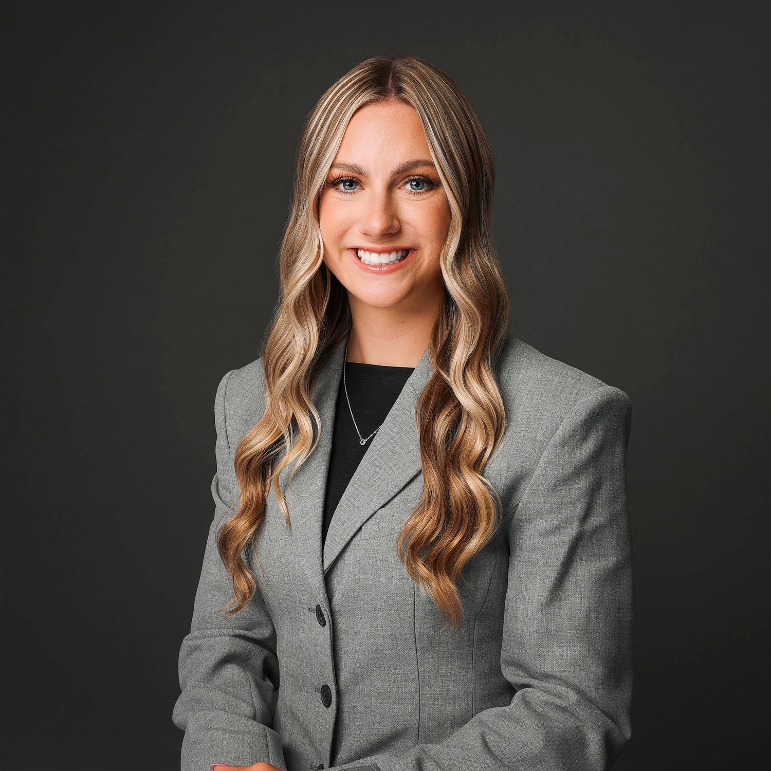 Professional headshot portrait of someone wearing a gray blazer against a dark background.