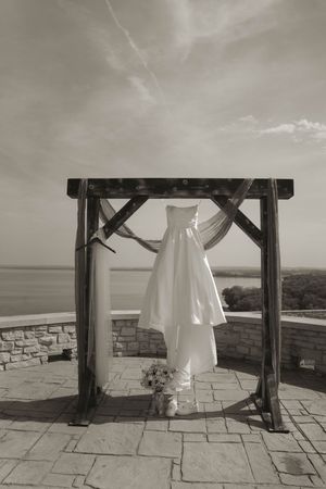 Black and white photo of wedding dress detail under wooden pergola with stone backdrop.