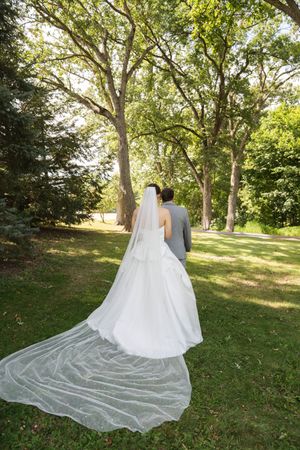 Wedding couple walking through a tree-lined path with a flowing cathedral veil trailing behind.