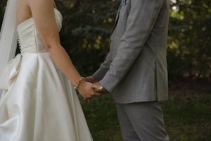 Close-up of couple holding hands during their wedding ceremony, bride in white dress and groom in grey suit.