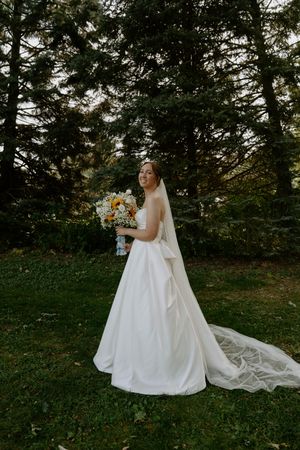 Elegant bridal portrait with sunflower bouquet against evergreen trees and flowing cathedral veil.