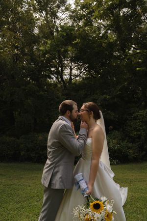 A romantic wedding moment captured outdoors with a sunflower bouquet and wooded backdrop.