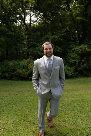 A groom in a light gray suit poses confidently on a grassy lawn with trees in the background.