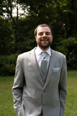 A joyful portrait of a groom in a gray suit and tie against a green forest backdrop.