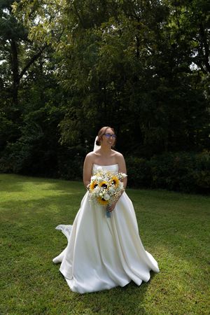 A bride in a flowing white dress holds a vibrant sunflower bouquet while standing on a lawn.
