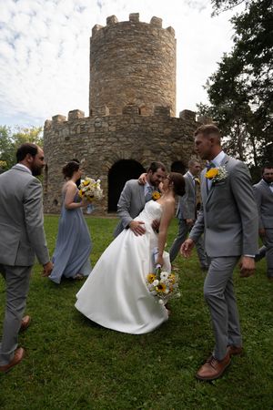 Joyful wedding party poses in front of a stone castle tower on grassy hill.