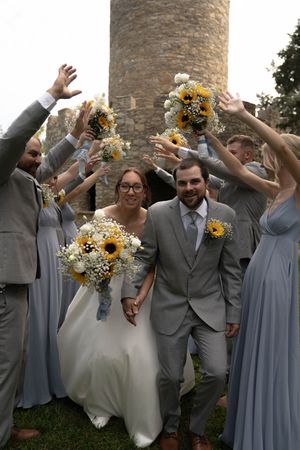 Wedding party celebrates with raised arms in front of historic stone tower.