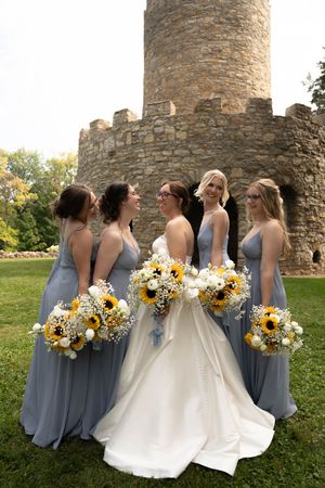 Wedding party in blue bridesmaid dresses holding sunflower bouquets pose near stone tower.