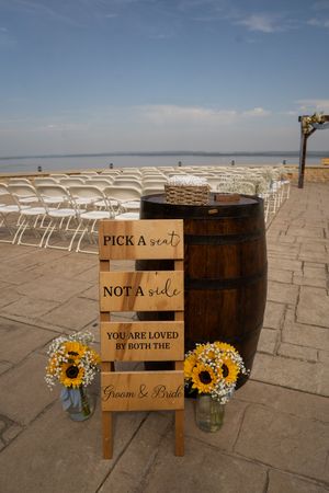 Rustic wedding ceremony decor with wooden barrels, sunflowers and seating sign.