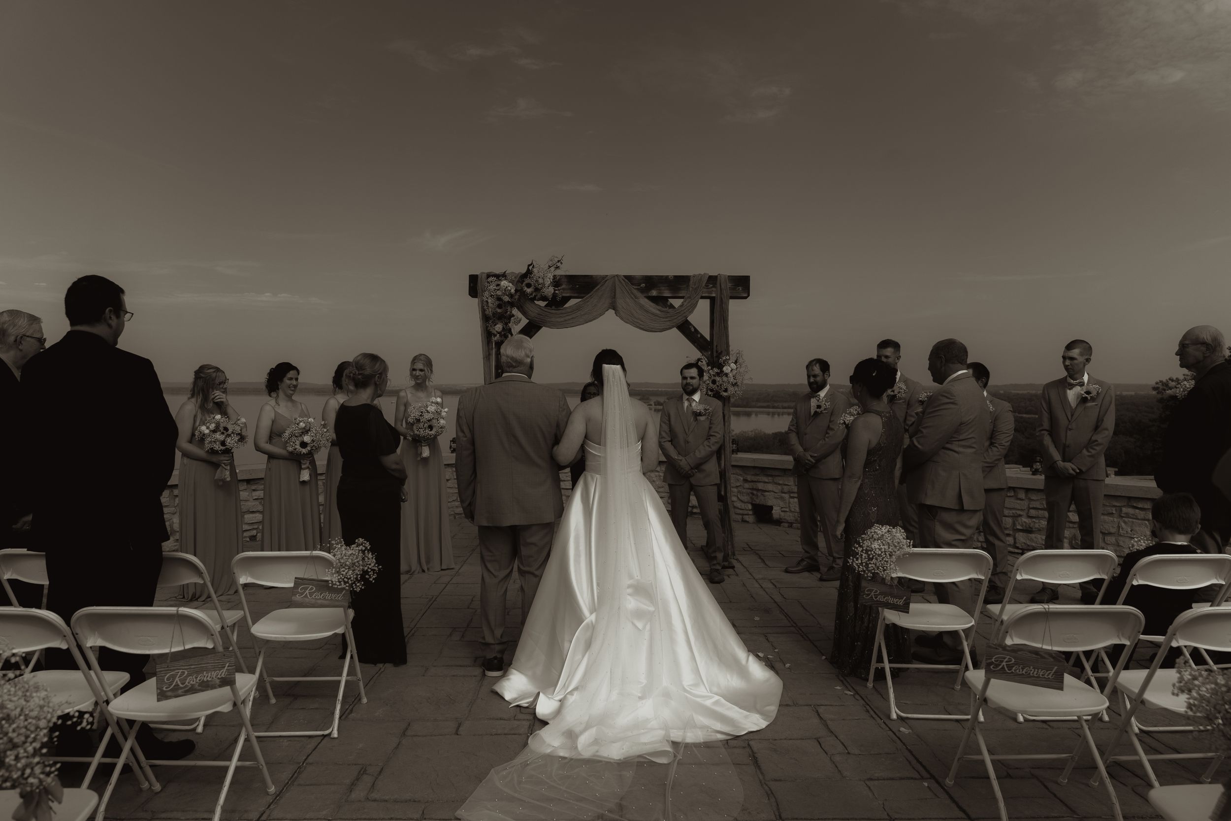 Black and white photo of outdoor wedding ceremony with guests seated at scenic venue.