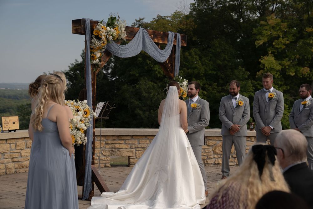 Wedding ceremony under decorated wooden arch with bridal party and guests at sunset.