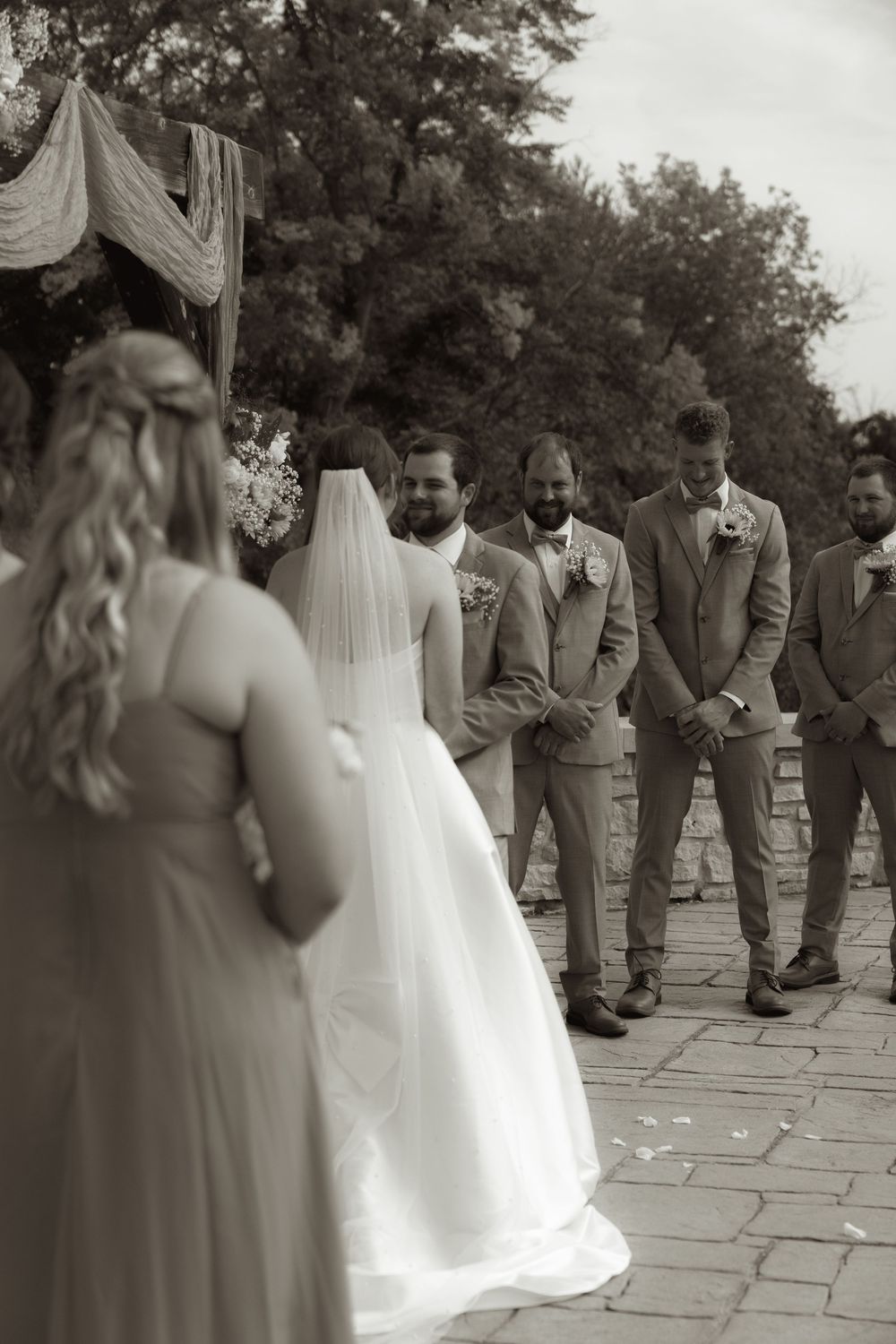 Black and white photo of wedding ceremony with bridal party standing at decorated outdoor altar.
