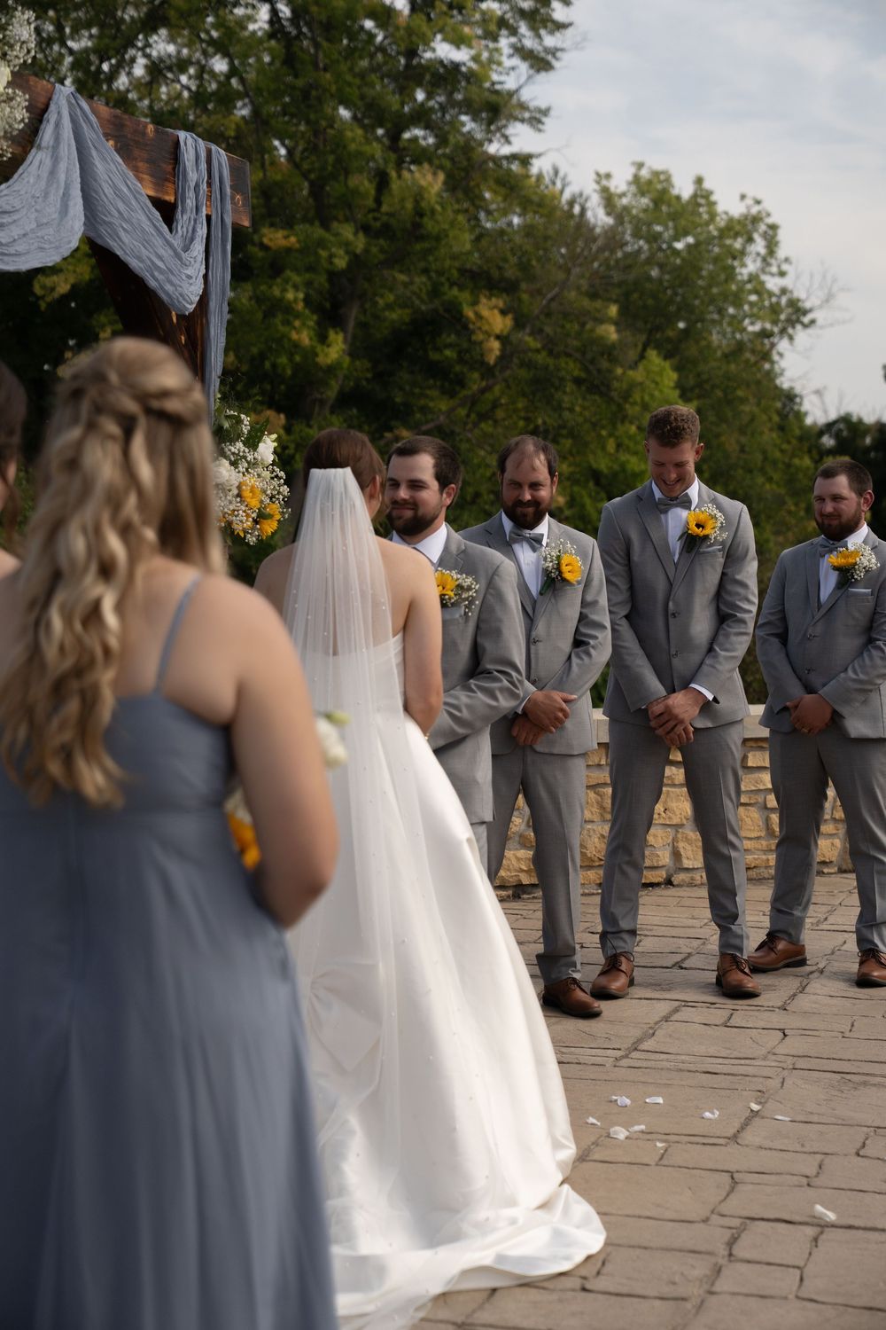 Wedding party in gray suits and blue dresses standing on stone patio during outdoor ceremony.