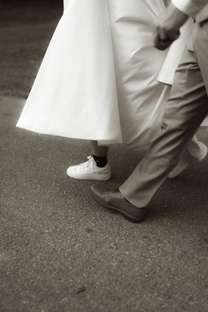 Black and white photo showing wedding shoes with a white dress hem and formal pants on a dark ground.