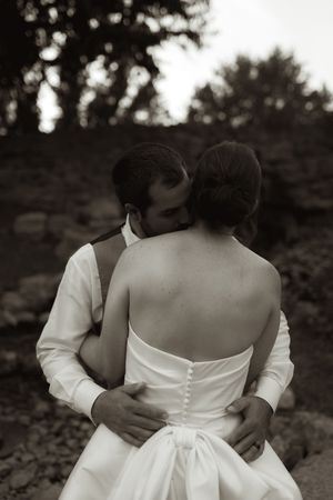 Back view of couple in wedding attire embracing against dark foliage.