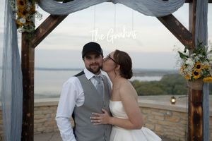 Wedding ceremony setup with rustic wooden arch decorated with flowers overlooking water at sunset.