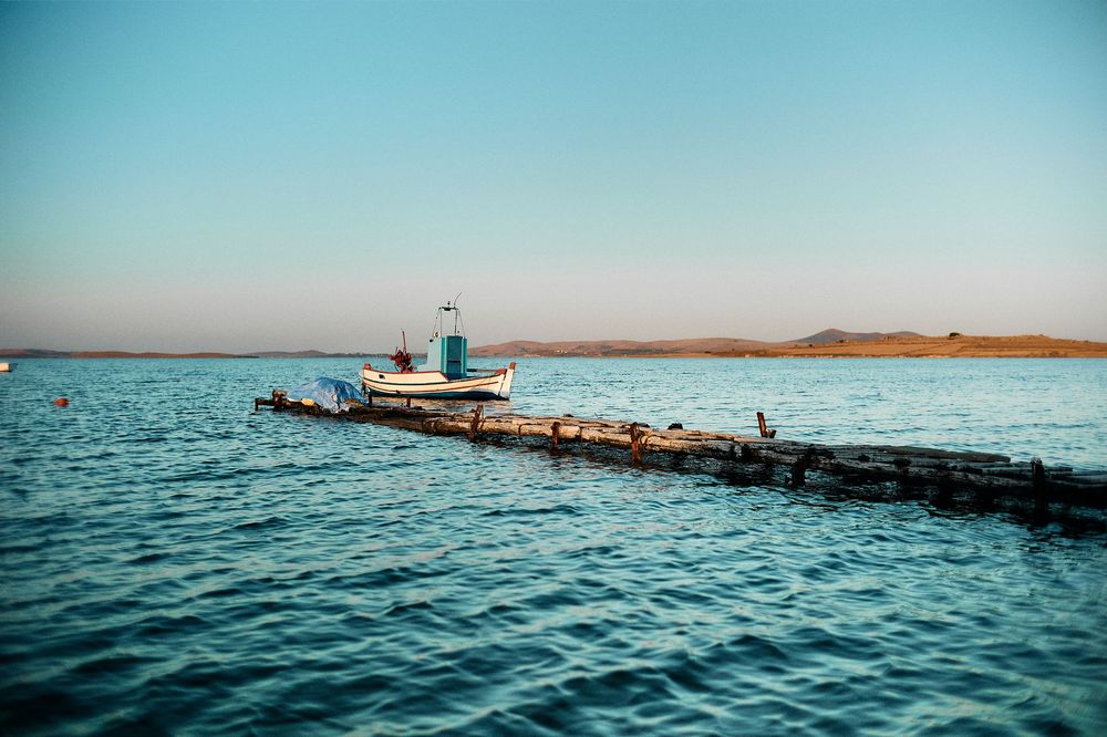 Small fishing boat cruises across tranquil blue waters near a wooden dock on a peaceful coastal morning.