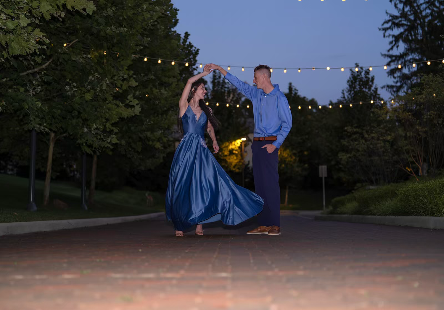 A couple dances under string lights at dusk in an outdoor setting.