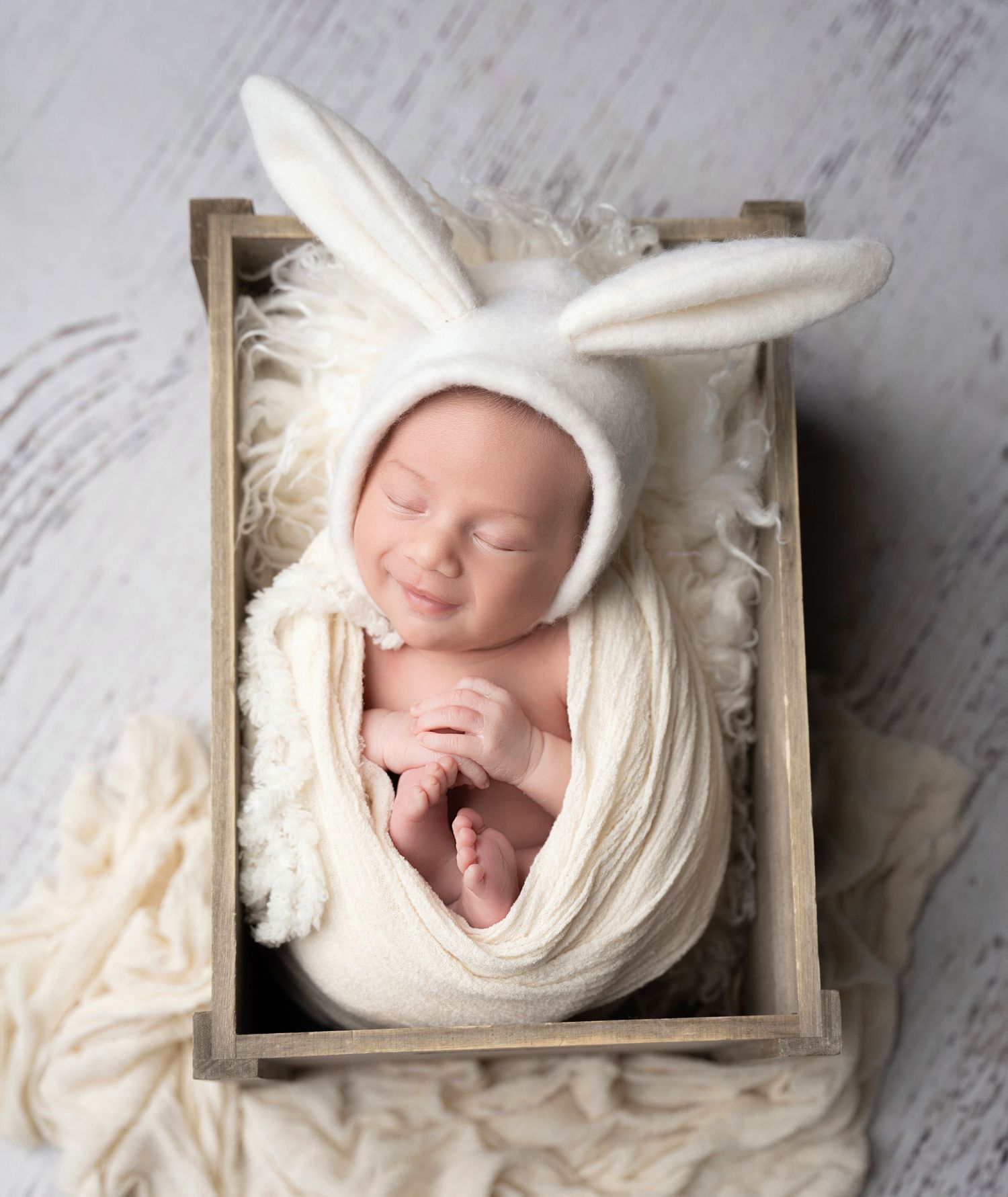 Miami Maternity photographer A sleeping newborn wearing a white bunny hat rests in a rustic wooden box surrounded by soft white fabric.
