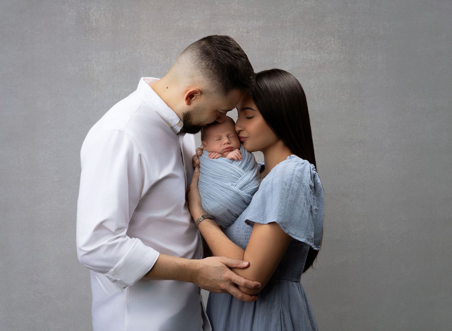 Miami Maternity photographer couple in light blue and white clothing share intimate moments in a series of tender poses against a gray wall.